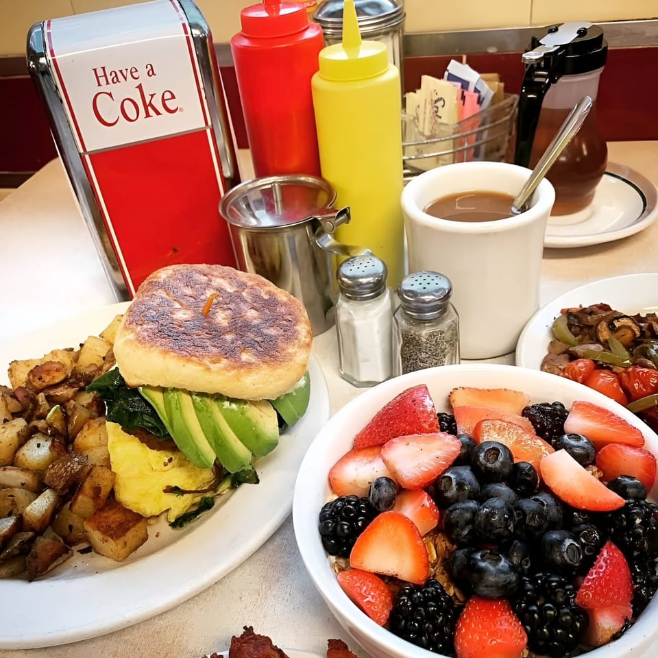 Diner breakfast with a sandwich, home fries, fruit bowl, coffee, and condiments on a table.