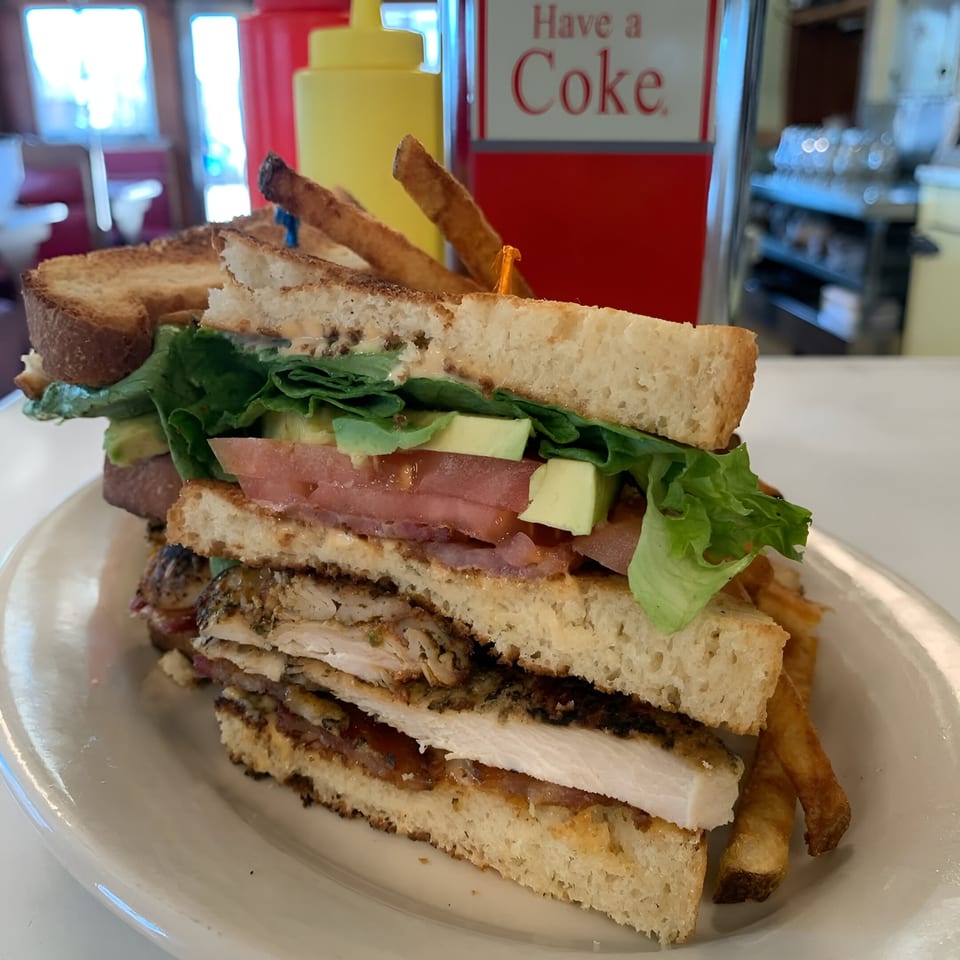 A stacked Parkway Diner club sandwich with fries, avocado, tomato, and lettuce on a white plate.