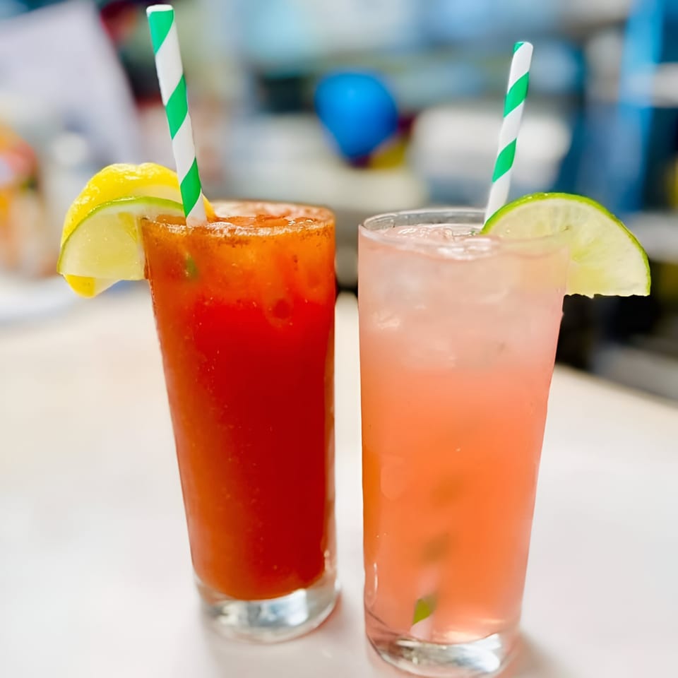 Two refreshing drinks, a red Bloody Mary with lemon and a pink lemonade with lime, on a diner counter.