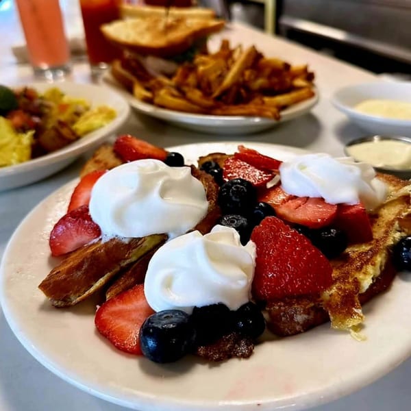 French toast topped with whipped cream, strawberries, and blueberries, with fries and drinks in the background.