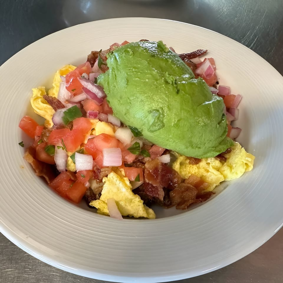 Scrambled eggs with bacon, diced tomatoes, onions, and a large scoop of avocado on a white plate.